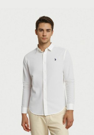 Jeune homme portant une chemise blanche à manches longues avec un petit logo sombre sur la poitrine et un pantalon beige, debout devant un fond uni.