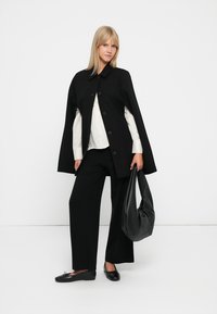 Cappa nera di lana con colletto alto e grandi bottoni, indossata sopra una maglia bianca e pantaloni larghi neri, completata da una grande borsa nera.