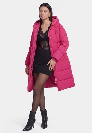 Frau trägt einen langen pinkfarbenen Steppmantel über einem schwarzen Spitzenoberteil, einem schwarzen Minirock, transparenten Strumpfhosen und schwarzen hochhackigen Stiefeletten und steht vor weißem Hintergrund.