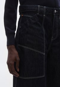 Traperice tamne boje s kontrastnim bijelim šavovima, velikim bočnim džepom i krojem koji pristaje u struku. Tkanina je teksturirana s glatkim završetkom.