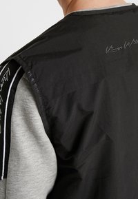 Svart ärmlös jacka med glänsande yta, med grå armhål och grå krage. Subtil vit broderad signaturdetalj på baksidan.