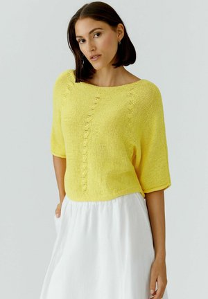 Frau mit kurzen dunklen Haaren trägt einen gelben kurzärmligen Strickpullover und einen weißen Rock, steht mit einer Hand in der Rocktasche.