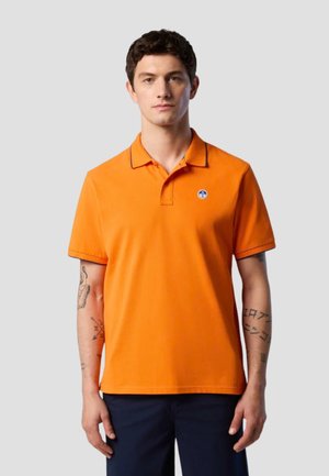 Giovane uomo con capelli corti e scuri, che indossa una polo arancione brillante e pantaloni scuri, in piedi davanti a uno sfondo grigio uniforme.