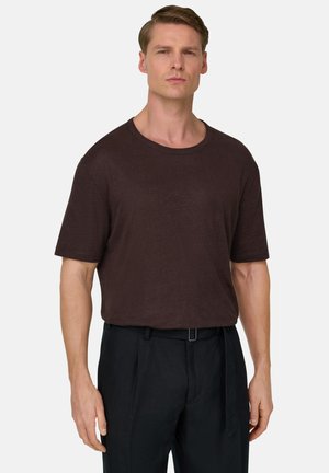 Homme aux cheveux châtain clair courts portant une chemise à manches courtes marron foncé et un pantalon noir avec une ceinture, sur un fond uni.