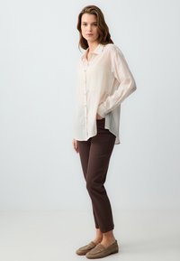Lichtroze overhemd met knoopjes en een iets langere achterkant, gecombineerd met bruin slim fit broek en beige instapchoenen. Minimalistisch ontwerp en zachte textuur.