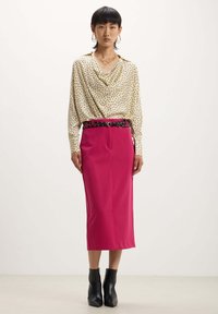 Blusa crema a pois con scollatura drappeggiata, abbinata a una gonna a tubino fucsia, cintura nera e stivaletti. Tessuti dalla consistenza liscia.