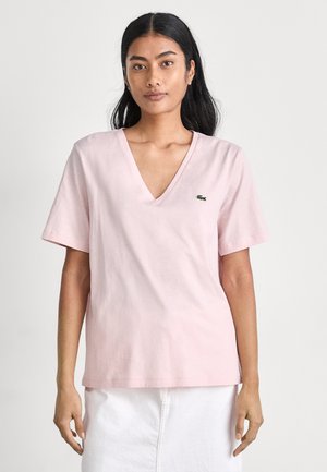 Pink cotton t-shirt with a v-neckline and short sleeves. Features a small green Lacoste logo on the chest. Soft texture and casual fit.