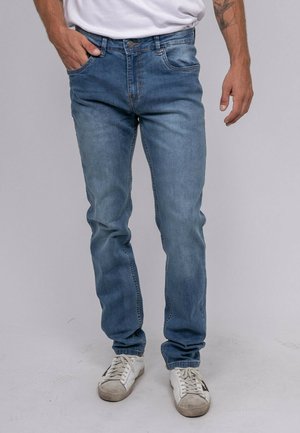 Homme portant un jean bleu clair, des baskets blanches et une chemise blanche, debout avec une main dans la poche devant un fond uni.