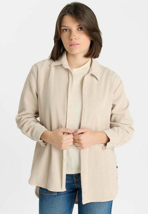 Jonge vrouw met steil bruin haar, een beige geribde jas, licht shirt en blauwe spijkerbroek dragend, staande tegen een effen achtergrond.
