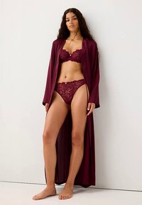 Accappatoio in raso color bordeaux con maniche lunghe, bralette in pizzo e bikini coordinato. L'accappatoio presenta un design fluido che mette in risalto i dettagli elaborati del pizzo.