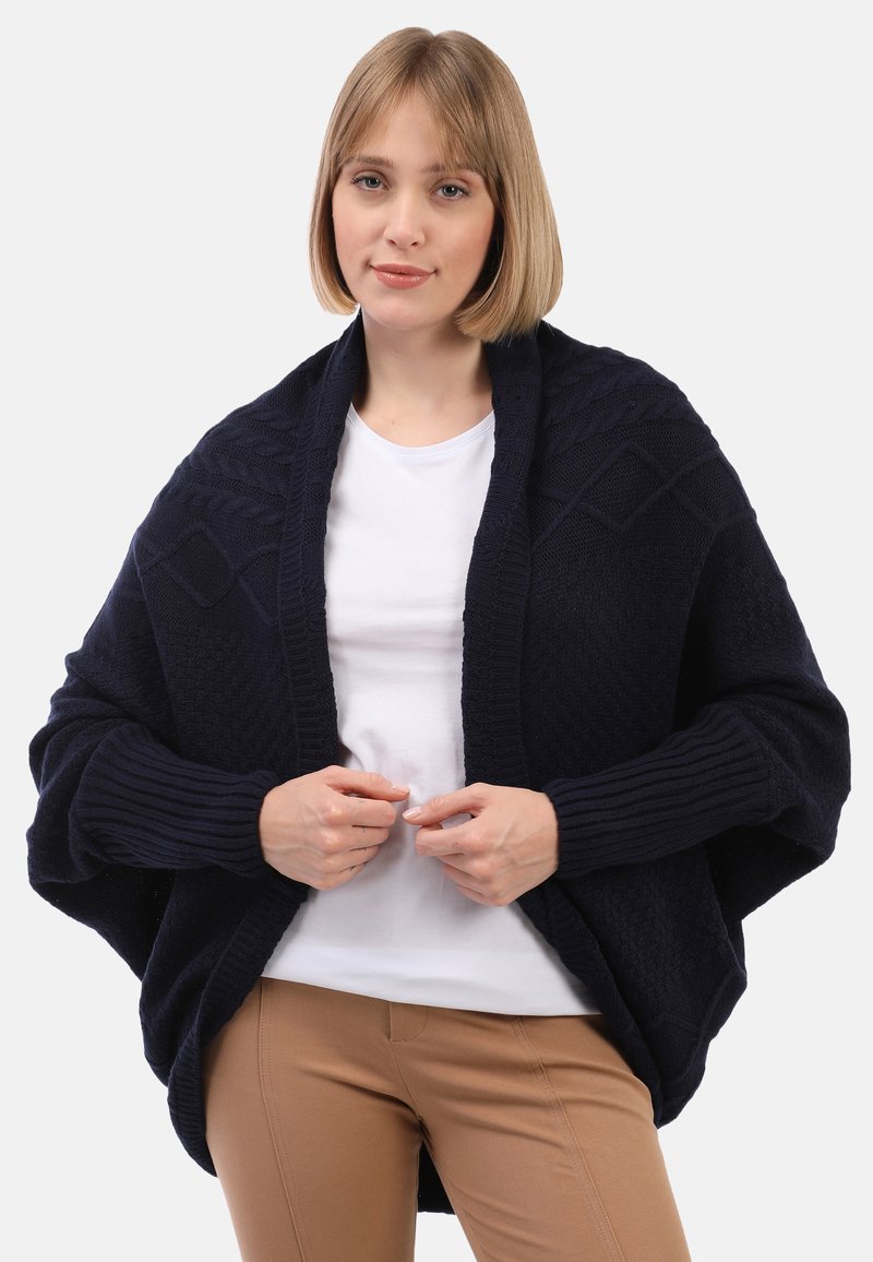 Mörkblå stickad sjal med diamantmönster och ribbade ärmar; bärs över en vit topp med en lös, oversized design.