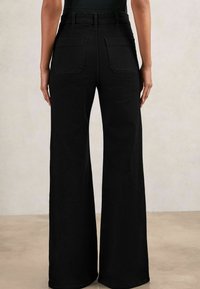 Persona viste pantalones de pierna ancha de talle alto en negro con dos bolsillos traseros, de pie contra un fondo neutro y liso.