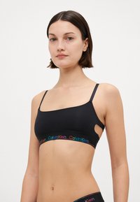 Svart bralette laget av mykt stoff. Den har tynne stropper, et utskåret design, og et flerfarget Calvin Klein-logo belte langs bunnen.