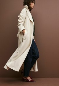 Palton lung bej din lână, cu rever oversized și buzunare frontale, asortat cu blugi bleu închis și toc red slingback. Textură netedă.