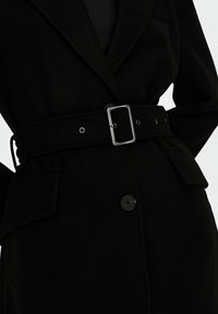 Manteau en laine noire avec taille ceinturée, grandes poches avant et une fermeture à un seul bouton. Texture douce avec un design structuré.