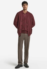 Burgunderfarbener Cardigan über einem gerippten Pullover, kombiniert mit hellbraunen Hosen und schwarzen Schuhen. Der Cardigan hat eine durchgehende Knopfleiste und Taschen.