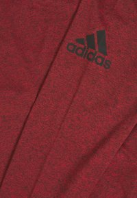 Punainen urheilupaita, joka on valmistettu teksturoidusta kankaasta ja jossa on näkyvä musta Adidas-logo edessä. Yksinkertainen muotoilu, lyhyet hihat.