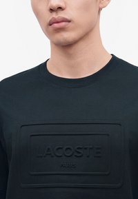 Κοντινό πλάνο ενός ατόμου που φοράει μαύρο t-shirt Lacoste Paris με ανάγλυφο ορθογώνιο λογότυπο στο στήθος.