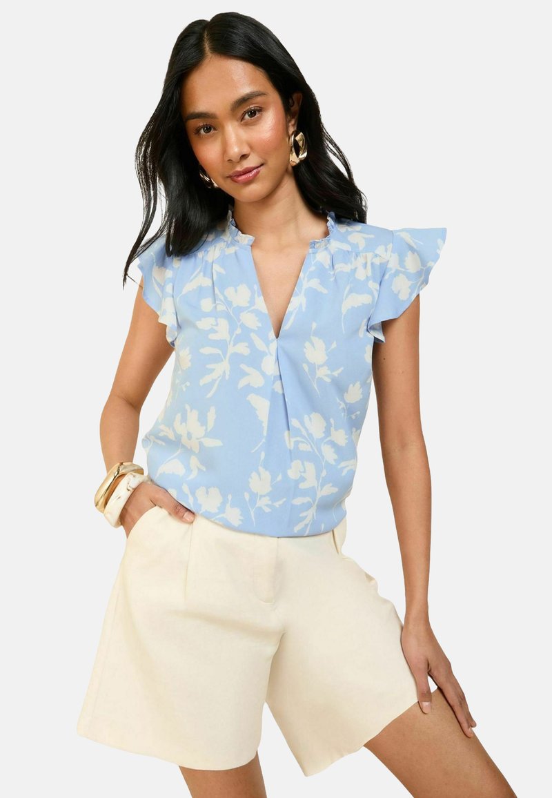 Blusa azzurra chiara con motivo floreale, maniche arricciate, scollatura a V, abbinata a pantaloni corte color crema. Il modello indossa orecchini a cerchio grandi e braccialetti impilati.