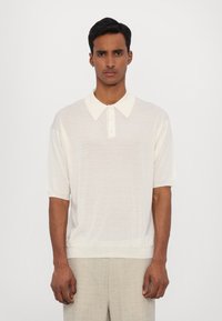 Polo shirt crème à manches courtes avec patte de boutonnage et col classique. Texture douce et coupe décontractée, associé à un pantalon gris clair.