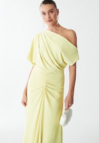 Robe jaune à une épaule avec un tissu drapé, présentant des fronces à la taille. Elle tient une pochette blanche avec un design texturé et ondulé.