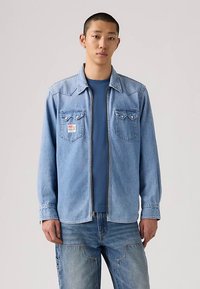 Νεαρός άνδρας με ανοιχτό μπλε πουκάμισο τζιν πάνω από σκούρο μπλε μπλουζάκι και μπλε τζιν, στέκεται μπροστά από απλό ανοιχτό φόντο.