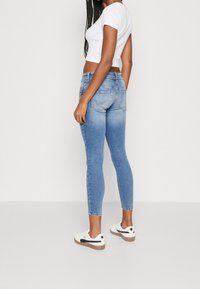 Jeans skinny azules con una textura desgastada, cintura alta y bolsillos traseros, combinados con una camiseta corta blanca y zapatillas blancas.