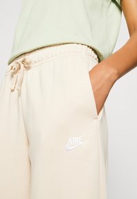 Beige sweatpants with an elastic waistband, drawstring, and pockets. Features a white embroidered Nike logo on the left thigh.