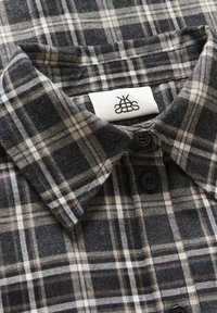 Dark gray plaid shirt collar with white and brown lines, featuring black buttons and a white label with a black abstract logo inside.