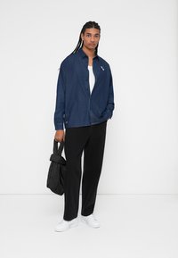 Denim button-up shirt in dark blue with a single white logo, paired with black loose-fitting pants and white sneakers, holding a black bag.