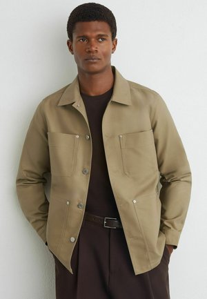 Giovane uomo che indossa una giacca beige sopra una camicia e pantaloni scuri, in piedi contro un muro bianco semplice con le mani nelle tasche.