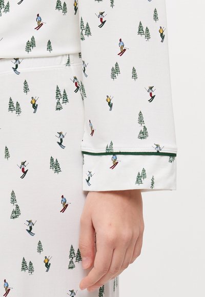 Σετ πιτζάμας με θέμα το σκι, σε λευκό ύφασμα, διακοσμημένο με πράσινα δέντρα και πολύχρωπους σκιέρ σε επαναλαμβανόμενο μοτίβο; η μανσέτα έχει πράσινη περιθώριο.