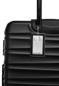 Valise rigide noire avec une surface texturée, des rainures verticales, une poignée rétractable et un porte-étiquette transparent attaché.