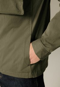 Eine Hand ruht in der Seitentasche einer olivgrünen Jacke mit sichtbaren Nähten und dunklen Jeans.