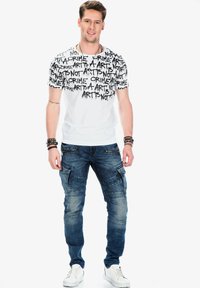 Witte T-shirt met zwart tekstpatroon met woorden zoals "KUNST" en "MISDAAD". Draagt een blauwe denim cargo broek en witte sneakers.