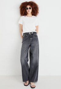 Bredde ben, falmede sorte jeans med en slidtegn tekstur, parret med en simpel hvid t-shirt og et sort nitterbælte. Flade sandaler fuldender looket.