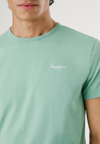 Mentazöld pamut póló, kerek nyakkivágással, fehér "Pepe Jeans" logóval a bal mellrészen. Sima textúra, rövid ujjú.