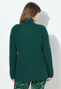 Mörkgrön, ribbad stickad tröja med hög krage och långa ärmar, kombinerad med gröna byxor med bladmönster. Texturerad tygdetalj synlig.