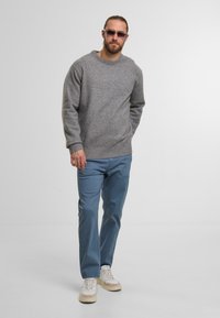 Maglione grigio con scollatura tonda, tessuto strutturato, abbinato a pantaloni azzurri chiari e sneakers beige. Gli occhiali da sole completano il look.