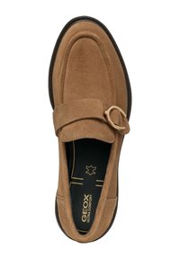 Mocasines de ante marrón con punta redonda, que presentan una tira decorativa y un acento de hardware circular. Suela negra con detalles de costura en contraste.