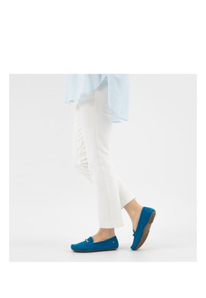 Persona che indossa pantaloni bianchi e mocassini blu con suola in gomma, che cammina su uno sfondo bianco uniforme.