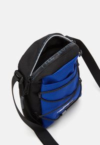 Crossbody torba iz črne in modre tkanine, s sshodnim zgornjim delom, več sprednjimi žepi in nastavljivim trakom. Lahka zasnova.