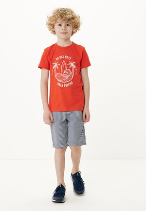 Rotes T-Shirt mit weißem Grafikdruck, gestreifte Shorts in Blau und Weiß sowie marineblaue Sneakers. Das Kind steht vor einem einfarbigen weißen Hintergrund.