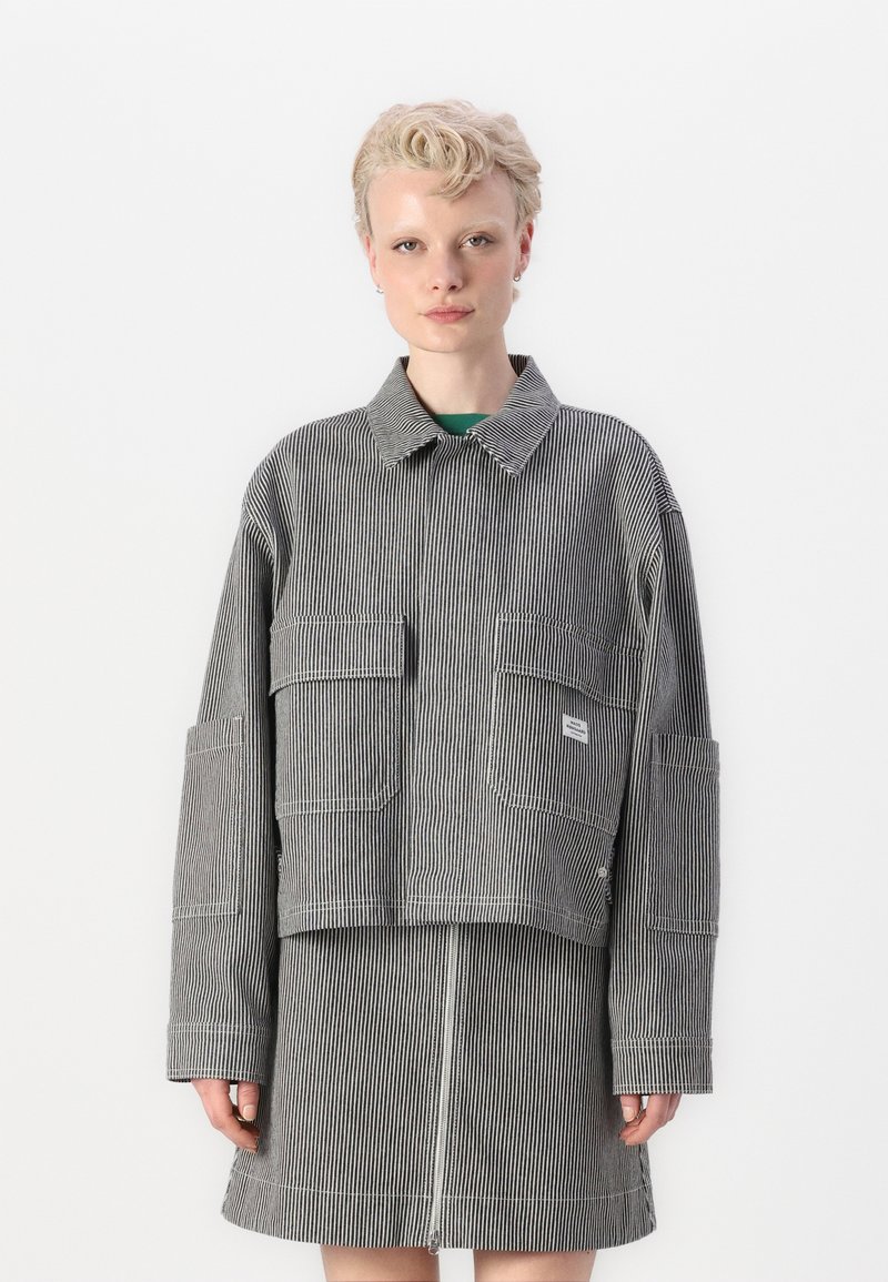 Chaqueta de mezclilla a rayas con cuello tipo polo y múltiples bolsillos frontales, combinada con una falda a juego que cuenta con un cierre de cremallera. Los colores son blanco y negro.