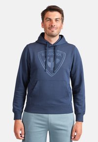 Tmavě modrá mikina s kapucí a klokaní kapsou. Má tónovaný logo motiv na hrudi a žebrované manžety a lem.