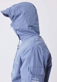 Giacca impermeabile blu chiaro con cappuccio e visiera corta, tasca con zip e logo sulla manica, e coulisse regolabile sul cappuccio.