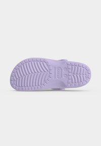 Η σόλα ενός ανοιχτόμωβ Crocs με ανάγλυφο σχέδιο και σημάδια μεγέθους μπροστά σε απλό ανοιχτό φόντο.