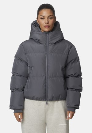 Vrouw draagt een donkergrijze gewatteerde jas met een hoge kraag en rits, gecombineerd met lichtgrijze joggingbroek tegen een effen achtergrond.