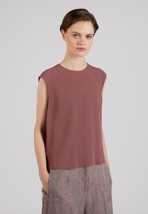 Femme aux cheveux courts et foncés portant un haut sans manches mauve et un pantalon gris texturé taille haute, debout avec les mains dans les poches, fond neutre.