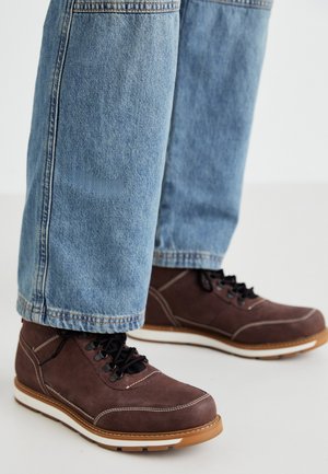 Zapatos de cuero marrón con cordones negros, costuras blancas y suela de goma. Combinados con unos jeans de mezclilla azul claro, de corte ligeramente holgado.